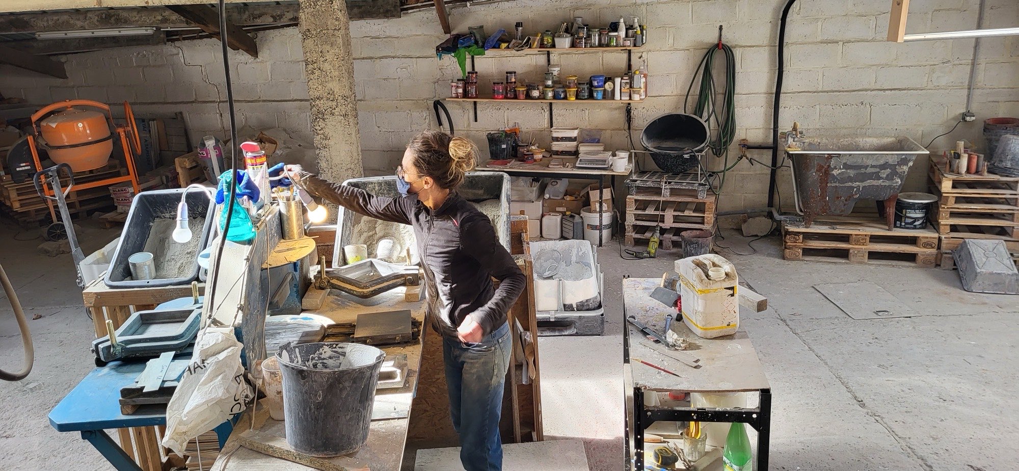 Intérieur de l'atelier : établi de moulage, bacs de ciment, pigments sur étagère et bétonnière, pendant la fabrication de carreaux