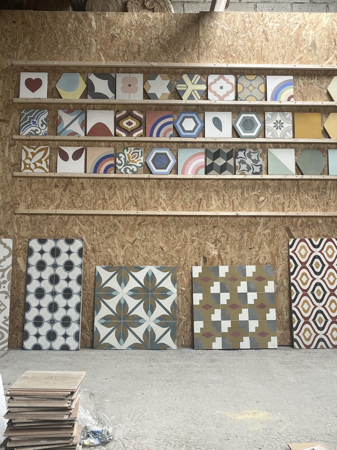 Mur de l'atelier : étagères de carreaux de ciment carrés et hexagonaux à motifs variés, et grands panneaux de carreaux assemblés posés au sol