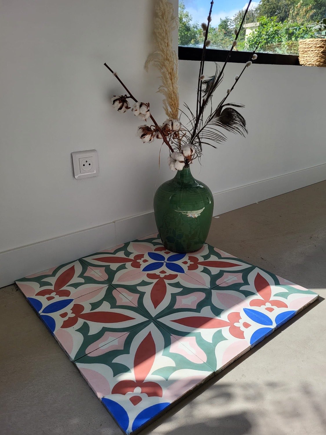 Carreaux de ciment à motif fleuri vert, rose et bleu posés au sol dans un angle de pièce, sous un vase en grès vert